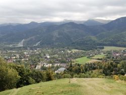 Zdjęcie przedstawia panoramę polskich Tatr, na pierwszym planie zielona łąka, w dole zabudowania, w tle Tatry i błękitne niebo. 