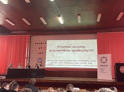 Na zdjęciu widać salę audytoryjną, w której odbywa się debata dotycząca przemocy. Na scenie, po lewej stronie, siedzą cztery osoby przy długim stole — wyglądają na prelegentów lub przedstawicieli instytucji biorących udział w spotkaniu.  W centralnej części znajduje się duży ekran z prezentacją zatytułowaną „Przemoc oczami pracowników społecznych!”. Pod tytułem widnieje lista trudnych zagadnień, z którymi mierzą się pracownicy socjalni, takich jak interwencje, problemy rodzinne, uzależnienia, agresja czy trudne warunki pracy.