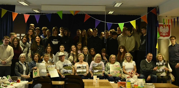 Na zdjęciu widać dużą grupę młodzieży i seniorów pozujących wspólnie w udekorowanej sali. Z przodu siedzą uczestnicy trzymający plakaty, a za nimi stoi grupa uczniów. Kolorowe girlandy i stoły z materiałami sugerują, że to było radosne wydarzenie warsztatowe lub integracyjne.