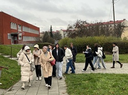 Na zdjęciu widać grupę osób – uczniów i seniorów – idących razem chodnikiem podczas spaceru. Większość z nich jest ciepło ubrana w kurtki, płaszcze i czapki, co wskazuje na chłodną, jesienno-zimową pogodę. W tle znajduje się czerwony budynek oraz blokowisko, a dookoła widoczna jest zieleń: trawniki i krzewy. Uczestnicy rozmawiają, śmieją się i wyglądają na zaangażowanych, co tworzy atmosferę integracji i wspólnego działania.