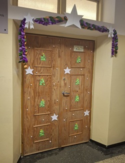 Na zdjęciu widać świątecznie udekorowane drzwi szkolnej sali. Drewniane drzwi ozdobione są papierowymi choinkami oraz małymi białymi gwiazdkami, rozmieszczonymi równomiernie na całej powierzchni. Nad drzwiami zawieszona jest kolorowa, skręcana girlanda w odcieniach zielonego, fioletowego i pomarańczowego. Girlandę podtrzymują duże, białe papierowe gwiazdy, które dodają dekoracji zimowego i odświętnego charakteru. Całość tworzy radosną, świąteczną atmosferę i wygląda jak element konkursu na najładniej udekorowane wejście do sali.