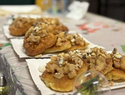 Na zdjęciu widać talerze z wypiekami ustawione w rzędzie na stole. Są to drożdżowe ciastka lub bułeczki z obfitą warstwą orzechów i kruszonki na wierzchu. Stół przykryty jest foliowym obrusem, a w tle widoczne są elementy świadczące o kiermaszu lub szkolnym wydarzeniu. Zdjęcie podkreśla domowy charakter wypieków i atmosferę wspólnego poczęstunku.