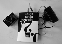 Na zdjęciu widoczna jest czarno-biała kompozycja. W centrum znajduje się album zatytułowany „Zapis socjologiczny. Tribute to Zofia Rydet”. Obok niego leży aparat fotograficzny oraz pokrowiec z paskiem. Całość nawiązuje do dokumentalnego charakteru fotografii i twórczości Zofii Rydet.