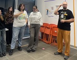 Na zdjęciu widać grupę osób biorących udział w warsztatach. Stoją w jasnym, nowoczesnym pomieszczeniu, a za nimi znajdują się pomarańczowe, składane krzesła ustawione w stos oraz przyklejone do ściany kartki z projektami lub szkicami. Po lewej stronie stoją trzy młode osoby – dwie z nich mają na sobie jasne, luźne bluzy i spodnie, a jedna ciemniejszą bluzkę w jasne kropki. Po prawej stronie stoi mężczyzna ubrany w czarną koszulkę z nadrukiem i pomarańczowe spodnie dresowe; wygląda na to, że coś wyjaśnia lub prezentuje, trzymając w ręku przedmiot przypominający marker lub wskaźnik.