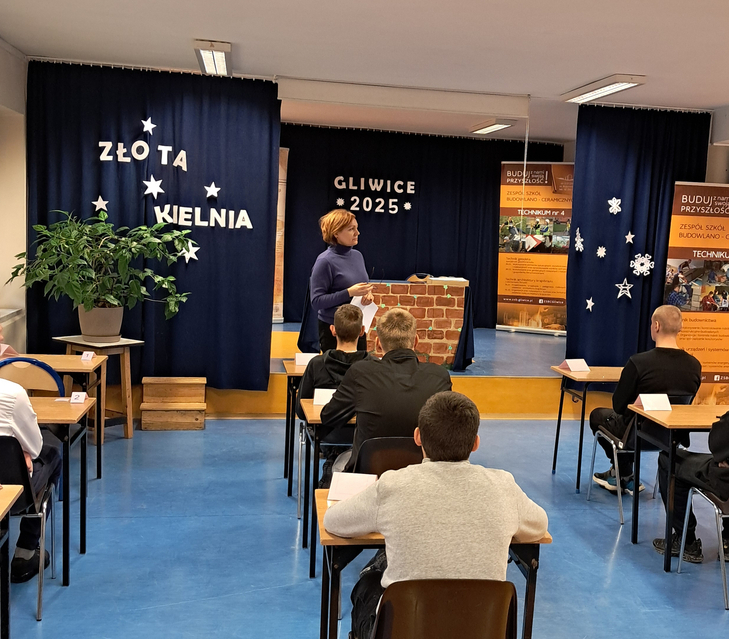Na zdjęciu widać salę szkolną podczas konkursu „Złota Kielnia” w Gliwicach 2025. Uczniowie siedzą w ławkach, a prowadząca stoi z przodu przy stanowisku z elementami murarskimi, obok banerów szkoły budowlanej.