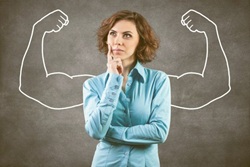 Na zdjęciu widać kobietę w niebieskiej koszuli, która wygląda na zamyśloną, a za nią narysowane są białą kredą umięśnione ramiona — symbol siły i pewności siebie.