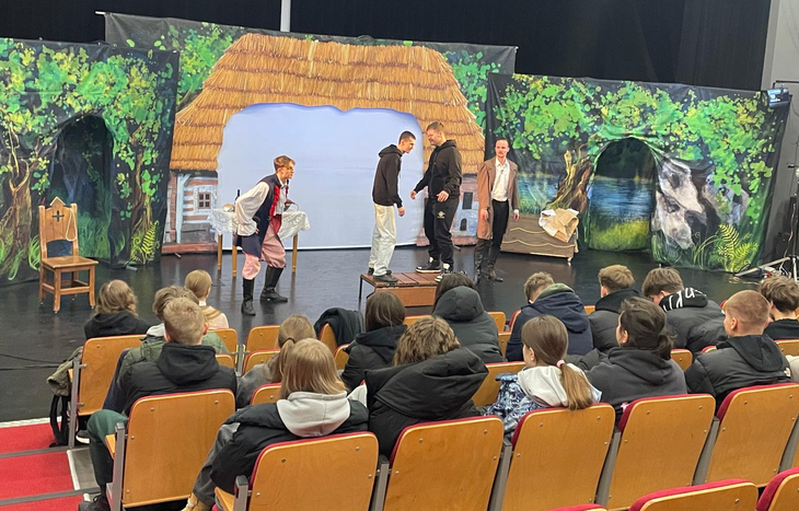 Na zdjęciu widać uczniów oglądających spektakl teatralny na scenie z bajkową scenografią, przedstawiającą chatę, las i jezioro, a aktorzy odgrywają scenę przed publicznością.