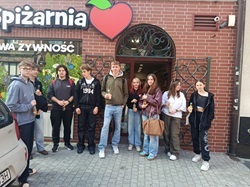 Na zdjęciu grupa uczniów klasy 5 AT pozuje przed wejściem do lokalnej eko Spiżarni. Uczniowie stoją razem przed budynkiem, uśmiechają się do zdjęcia i prezentują pamiątkową fotografię z ekologiczno-edukacyjnego spaceru po Gliwicach. Zdjęcie oddaje atmosferę wspólnego odkrywania miejsc związanych ze świadomą konsumpcją i ideą less waste.