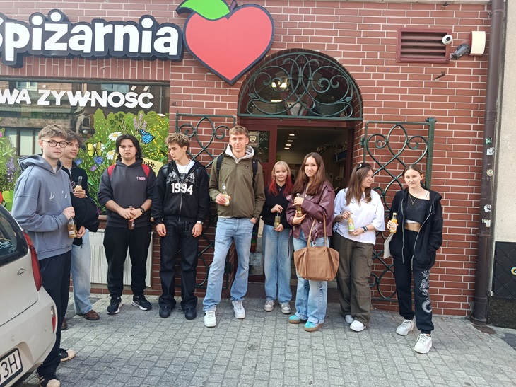 Na zdjęciu grupa uczniów klasy 5 AT pozuje przed wejściem do lokalnej eko Spiżarni. Uczniowie stoją razem przed budynkiem, uśmiechają się do zdjęcia i prezentują pamiątkową fotografię z ekologiczno-edukacyjnego spaceru po Gliwicach. Zdjęcie oddaje atmosferę wspólnego odkrywania miejsc związanych ze świadomą konsumpcją i ideą less waste.