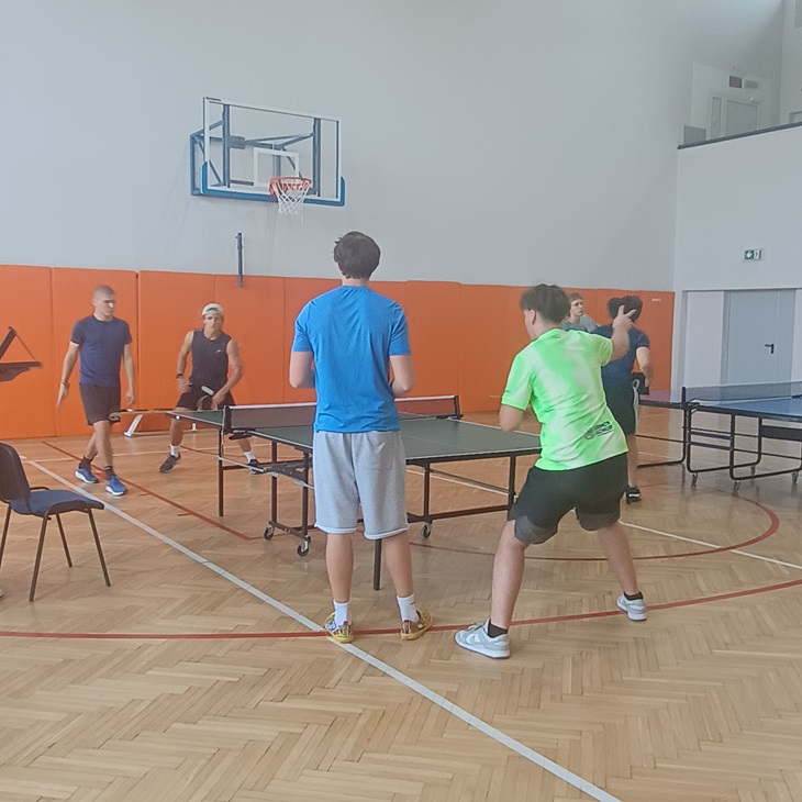 Na zdjęciu widać zawodników podczas rozgrywek tenisa stołowego. Mecz odbywa się w szkolnej hali sportowej, przy dwóch stołach. Zawodnicy są w trakcie dynamicznej wymiany, a w tle widoczni są pozostali uczestnicy turnieju. Fotografia dobrze oddaje sportową atmosferę, zaangażowanie i rywalizację towarzyszącą zawodom.