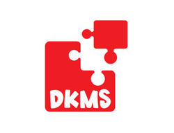 Na zdjęciu widać czerwone logo DKMS, przedstawiające dwa elementy puzzli symbolizujące dopasowanie dawcy i biorcy komórek macierzystych. Pod symbolem znajduje się napis „DKMS"