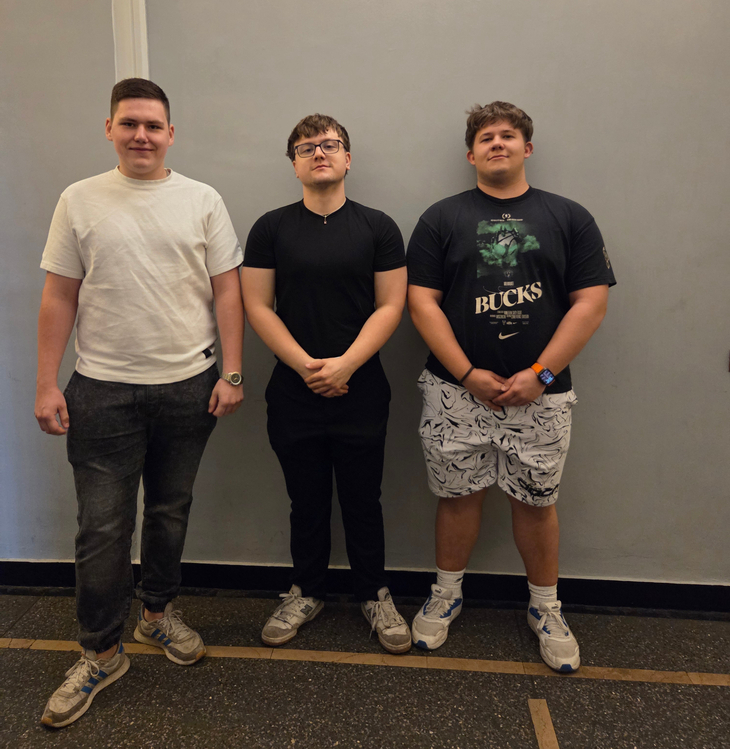 Na zdjęciu widać trzech uczniów stojących obok siebie na tle jasnej ściany w szkolnym korytarzu. Zdjęcie zostało wykonane podczas olimpiady, w której uczestnicy rozwiązywali zadania sprawdzające wiedzę z zakresu elektrotechniki, energetyki oraz zagadnień związanych z nowoczesnymi technologiami elektrycznymi i energetycznymi. Uczniowie stoją w swobodnej pozycji i pozują do wspólnego zdjęcia.