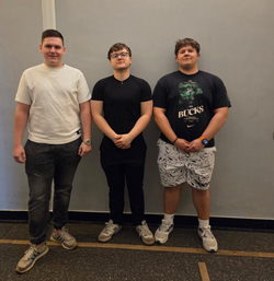 Na zdjęciu widać trzech uczniów stojących obok siebie na tle jasnej ściany w szkolnym korytarzu. Zdjęcie zostało wykonane podczas olimpiady, w której uczestnicy rozwiązywali zadania sprawdzające wiedzę z zakresu elektrotechniki, energetyki oraz zagadnień związanych z nowoczesnymi technologiami elektrycznymi i energetycznymi. Uczniowie stoją w swobodnej pozycji i pozują do wspólnego zdjęcia.