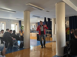 Na zdjęciu widać mężczyznę prowadzącego spotkanie w sali szkolnej. Stoi na środku pomieszczenia i przemawia do grupy uczniów siedzących po bokach. Uczestnicy uważnie go słuchają, a cała scena ma charakter edukacyjnego spotkania lub prelekcji.