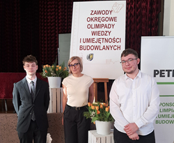 Na zdjęciu widać trzy osoby stojące przed tablicą z napisem „Zawody Okręgowe Olimpiady Wiedzy i Umiejętności Budowlanych”. Po bokach ustawione są dekoracje z kwiatami w donicach. Dwie osoby stoją po lewej i prawej stronie tablicy, a jedna znajduje się pośrodku. W tle widać czerwone zasłony oraz baner sponsora wydarzenia.