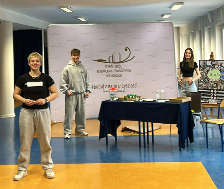 Na zdjęciu widać troje uczniów stojących w szkolnym holu przy stoisku promocyjnym Zespół Szkół Budowlano-Ceramicznych w Gliwicach. Za nimi znajduje się roll-up z logo szkoły i hasłem „buduj z nami przyszłość”, a obok stolik z materiałami informacyjnymi. Uczniowie uśmiechają się i wyglądają na zaangażowanych w prezentację.