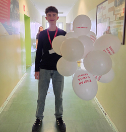 Na zdjęciu widać młodego chłopaka stojącego na szkolnym korytarzu. Ubrany jest w ciemną bluzkę z długim rękawem i jasne jeansy. Na szyi ma identyfikator na czerwonej smyczy. Trzyma w ręce duży bukiet białych balonów, na których widnieją czerwone napisy, m.in. „Zostań dawcą szpiku” oraz adres strony „www.dkms.pl”