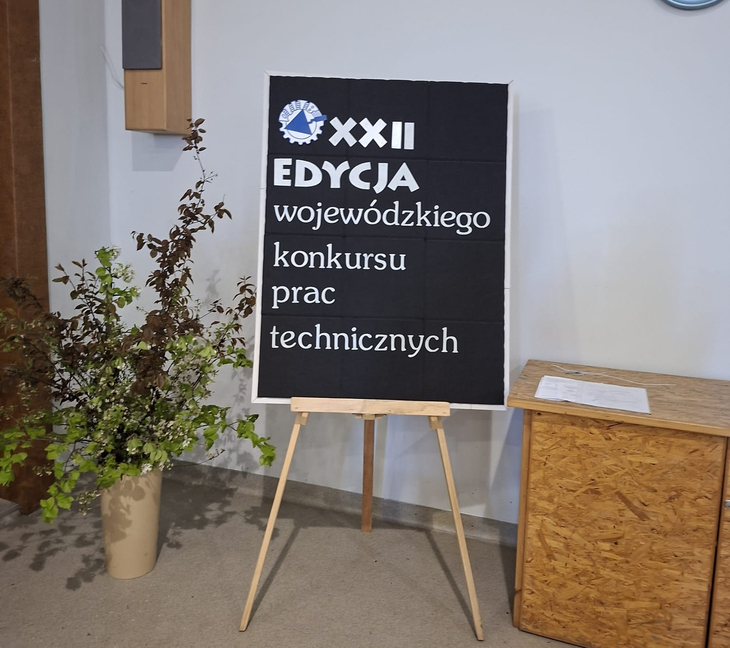 Na zdjęciu widać tablicę informacyjną ustawioną na sztaludze z napisem „XXII edycja wojewódzkiego konkursu prac technicznych”. Obok znajduje się stolik oraz dekoracyjna roślina