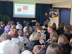 Na zdjęciu widać uczniów zgromadzonych na sali podczas prelekcji o cyberbezpieczeństwie. Prowadzący stoi przed ekranem z prezentacją, omawiając zagadnienia związane z zagrożeniami w sieci, a uczestnicy uważnie słuchają.