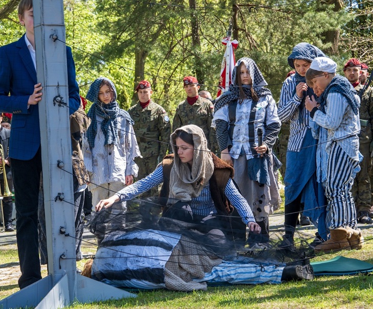 Na pierwszym zdjęciu widać młodzież biorącą udział w inscenizacji historycznej – ubrani w stroje stylizowane na więźniów obozów koncentracyjnych, otaczają symboliczną scenę przy ogrodzeniu z drutu kolczastego, oddając hołd ofiarom. W tle stoją również żołnierze, co podkreśla uroczysty i patriotyczny charakter wydarzenia.