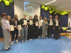 Na zdjęciu widoczna jest grupa uczniów wraz z nauczycielami podczas uroczystości zakończenia szkoły. Uczniowie stoją w jednym rzędzie, trzymając w rękach świadectwa lub dyplomy, co podkreśla rangę wydarzenia. Są elegancko ubrani – w koszule, marynarki i stroje galowe, co wskazuje na oficjalny charakter uroczystości.