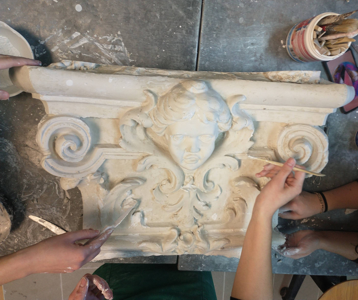Na zdjęciu widać ozdobny gipsowy element z twarzą i ornamentami. Kilka osób pracuje nad nim, wygładzając detale narzędziami.