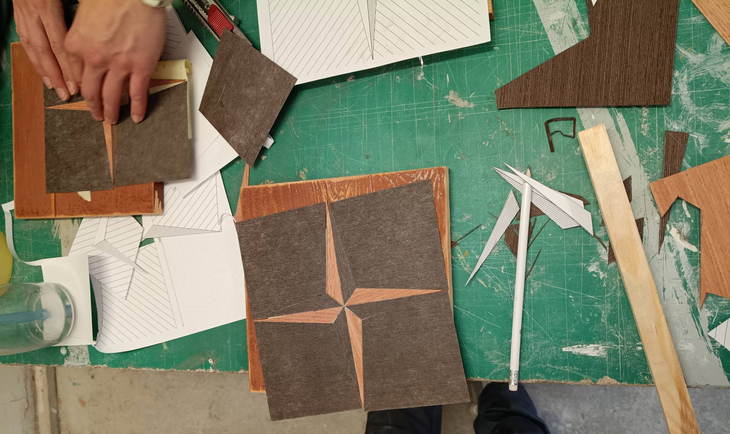 Na zdjęciu widać pracę warsztatową związaną z intarsją. Na stole leżą kawałki forniru i narzędzia, a uczniowie układają geometryczny wzór z różnych rodzajów drewna na podkładzie. Obok znajdują się szkice oraz materiały pomocnicze, co wskazuje na etap projektowania i precyzyjnego dopasowywania elementów.