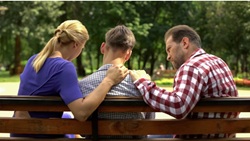 Na zdjęciu widać nastolatka siedzącego na ławce w parku, otoczonego przez dwoje dorosłych – prawdopodobnie rodziców. Oboje kładą mu ręce na plecach w geście wsparcia i troski, co sugeruje rozmowę w trudnym momencie i okazywanie emocjonalnej bliskości.