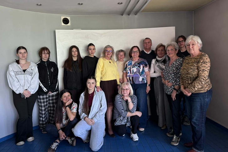 Na zdjęciu widać grupę młodzieży i seniorów pozujących razem w pomieszczeniu – stoją i siedzą obok siebie, uśmiechnięci, co sugeruje miłą i integracyjną atmosferę spotkania międzypokoleniowego.