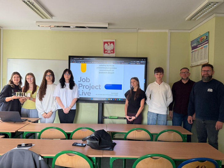 Na zdjęciu uczniowie wraz z nauczycielami prezentują projekt podczas szkolnego wydarzenia edukacyjnego. W sali lekcyjnej, przed ekranem z prezentacją „Job Project Live”, uczestnicy wspólnie przedstawiają efekty swojej pracy i rozwijają umiejętności zawodowe oraz językowe.