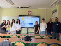 Na zdjęciu uczniowie wraz z nauczycielami prezentują projekt podczas szkolnego wydarzenia edukacyjnego. W sali lekcyjnej, przed ekranem z prezentacją „Job Project Live”, uczestnicy wspólnie przedstawiają efekty swojej pracy i rozwijają umiejętności zawodowe oraz językowe.