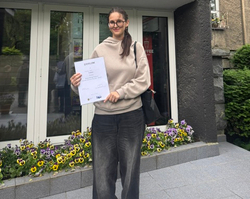 Na zdjęciu widoczna jest uczennica stojąca przed siedzibą Regionalnego Ośrodka Doskonalenia Nauczycieli „WOM” w Katowicach. W dłoniach trzyma dyplom potwierdzający udział w finale Wojewódzkiego Konkursu Wiedzy o Kulturze Fizycznej i Zdrowiu. Fotografia dokumentuje jej sukces oraz reprezentowanie szkoły w gronie finalistów konkursu.