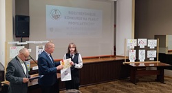 Na zdjęciu widać uroczystość wręczenia nagród podczas konkursu plastycznego. W centrum stoi młoda osoba odbierająca dyplom i torbę z upominkami od mężczyzny w garniturze. Obok, po lewej stronie, znajduje się drugi mężczyzna, który trzyma dokumenty i prawdopodobnie prowadzi ceremonię. W tle widoczny jest ekran projekcyjny z napisem w języku polskim: „Rozstrzygnięcie konkursu na plakat profilaktyczny…”, co sugeruje, że wydarzenie dotyczyło konkursu związanego z profilaktyką zdrowotną lub społeczną. Po obu stronach sceny ustawiono plansze z wyeksponowanymi pracami konkursowymi – rysunkami i plakatami wykonanymi przez uczestników. Wydarzenie odbywa się w sali konferencyjnej lub domu kultury. Atmosfera jest oficjalna i uroczysta, a zdjęcie dokumentuje moment wręczenia nagrody jednemu z laureatów konkursu.