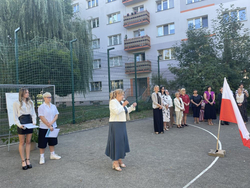 Uroczyste rozpoczęcie roku szkolnego na szkolnym boisku. W centrum dyrektor przemawia do uczniów i grona pedagogicznego, obok stoi polska flaga.