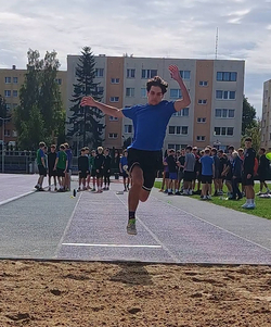 Zawodnik podczas skoku w dal. W tle widać pozostałych uczestników zawodów oraz osoby sędziujące konkurencję. Dynamiczne ujęcie pokazuje moment lotu nad piaskownicą.
