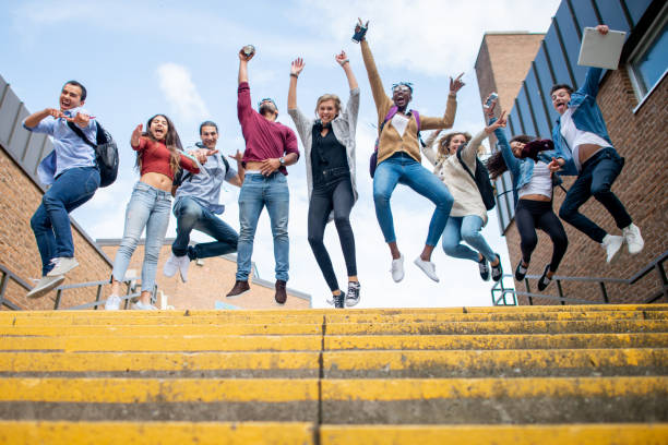 A diverse group of students at a top of a flight of stairs outside a school building. They are jumping mid-air and cheering on the last day of school.